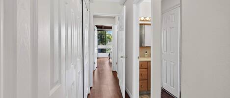 Un pasillo con puertas blancas, suelo de madera y vistas a una habitación con ventana y plantas.
