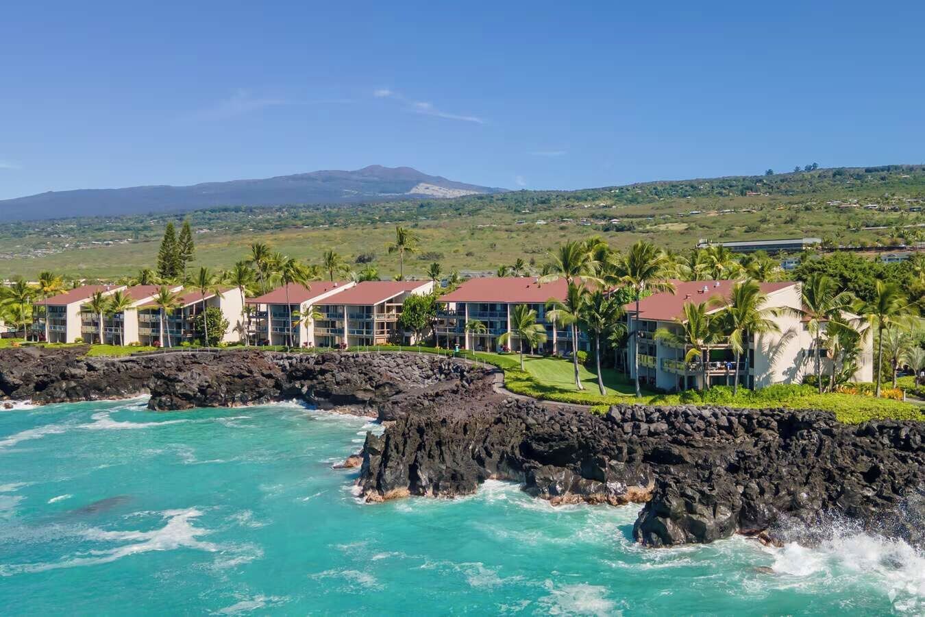 A coastal hotel with multiple buildings, a rocky shoreline, and a mountain in the background.