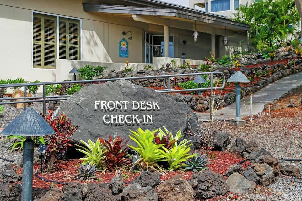 A front desk sign with 'FRONT DESK CHECK-IN' written on it, surrounded by rocks and plants.