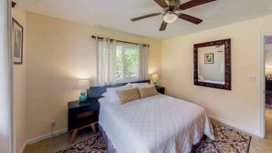 Un dormitorio con cama, mesitas de noche, ventilador de techo, ventana con cortinas, espejo y alfombra estampada.
