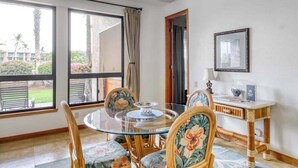 Una habitació amb una taula rodona de vidre, cadires florals, una taula auxiliar de fusta amb una làmpada i un quadre al mur.
