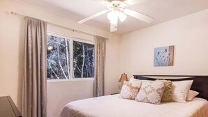 Um quarto com uma cama, ventilador de teto, janela com cortinas e um quadro na parede.