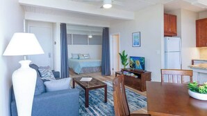 Een moderne woonkamer met een bank, salontafel en televisie, een afzonderlijke slaapkamer met een bed, en een keuken met kasten en een koelkast.
