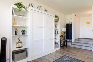Ein Zimmer mit einem weißen Kleiderschrank, Regalen mit Dekoration, einem kleinen Tisch mit einer Pflanze und einer Treppe, die in ein weiteres Zimmer führt.