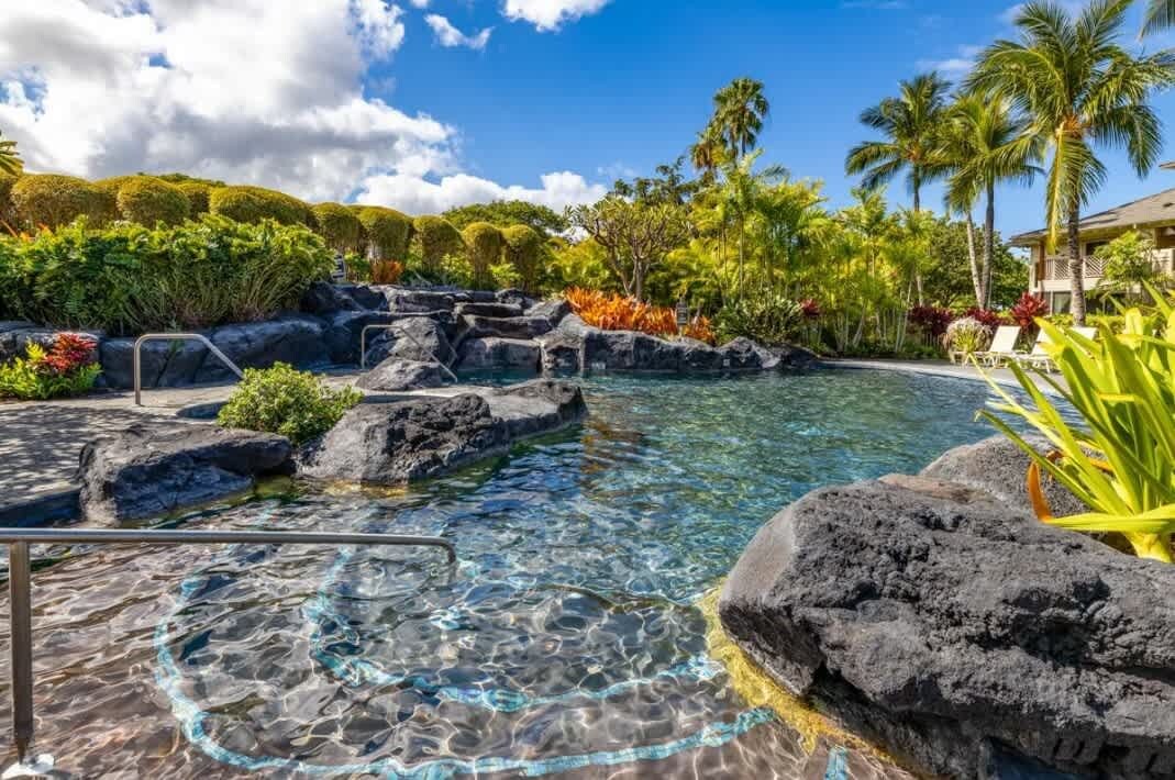 A clear pool with rock formations, surrounded by tropical vegetation and palm trees.