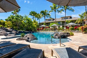 A pool area with lounge chairs, a small waterfall feature, and a building with balconies in the background. - Waikoloa Beach/Golf Resort Villa- Pool, Hot Tub, AC, Kitchen, Walk to Shops/Dine (Waikoloa)