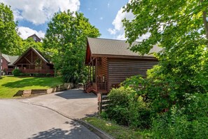 A log cabin with a porch, surrounded by lush greenery and a clear sky. - Bear Essentials Lodge (Sevierville)