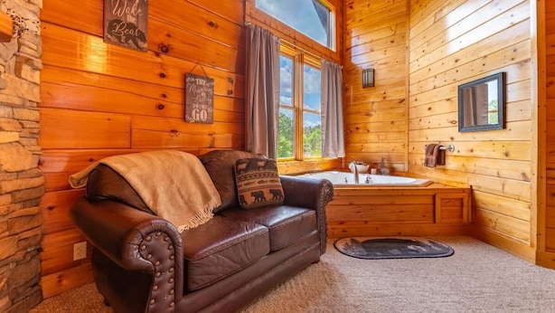 Interior acogedor de una cabaña con un sofá de cuero, pared de piedra, ambiente rústico de madera, ventana con cortinas y un baño con bañera.