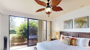Habitación de hotel con cama, ventilador de techo, mesita de noche con lámpara y vista a zonas verdes.