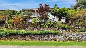 A house with a stone retaining wall, lush vegetation, and a white building with a balcony in the background. - NEW Kona Family Home and Ohana with Bay Sunsets (Kailua-Kona)