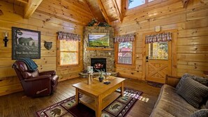 A log cabin interior with a stone fireplace, leather recliner, wooden table, and a television mounted above it. - Bear Hug (Pigeon Forge)