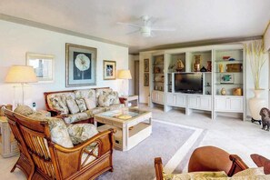 Sala de estar com sofá, poltronas, mesa de centro, televisão e prateleiras com objetos decorativos.
