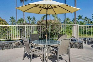 An outdoor patio with a round glass table and four chairs, an umbrella, and a white metal fence. - Keauhou Golf View Condo- Pool, Hot Tub, Kitchen, Laundry, BBQ, Lanai, Near Beach (Kailua-Kona)
