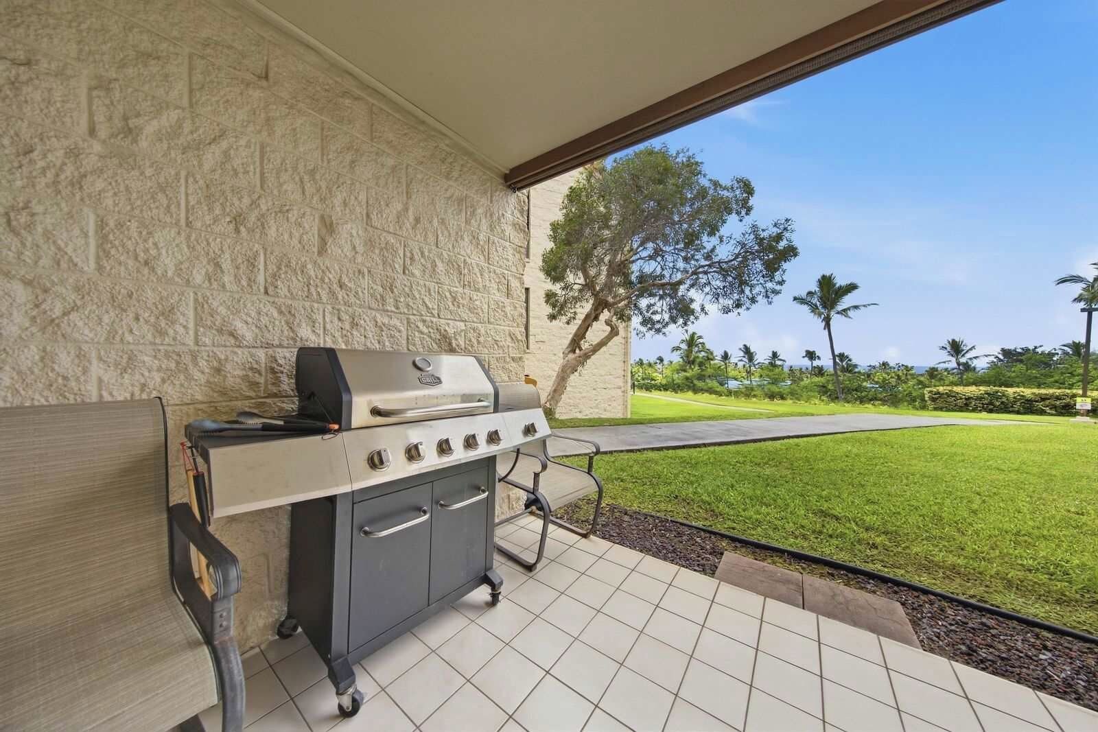 An outdoor grill area with a stone wall backdrop and a view of a well-maintained lawn and palm trees.