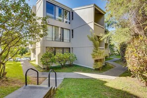 Um edifício residencial moderno de vários andares, com um gramado bem cuidado, um caminho que leva à entrada e uma pequena área de jardim com arbustos.