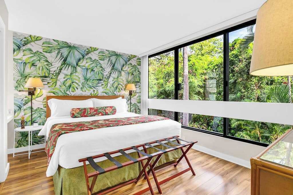 Un dormitorio con cama, un banco, una mesita de noche, una lámpara y vistas a zonas verdes a través de amplios ventanales.