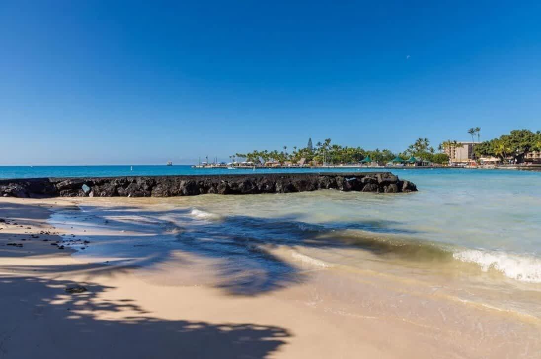 A beach with clear water, a rocky breakwater, and a distant shoreline with buildings and palm trees.