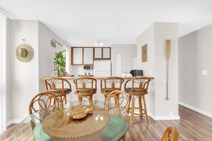 A dining area with a glass table and wicker chairs, a kitchen with white cabinets, and a wooden floor. - Kailua Bay Casa Tropical Kona Condo- Pool, Hot Tub, AC, WD- Walk to Beach Fun (Kailua-Kona)
