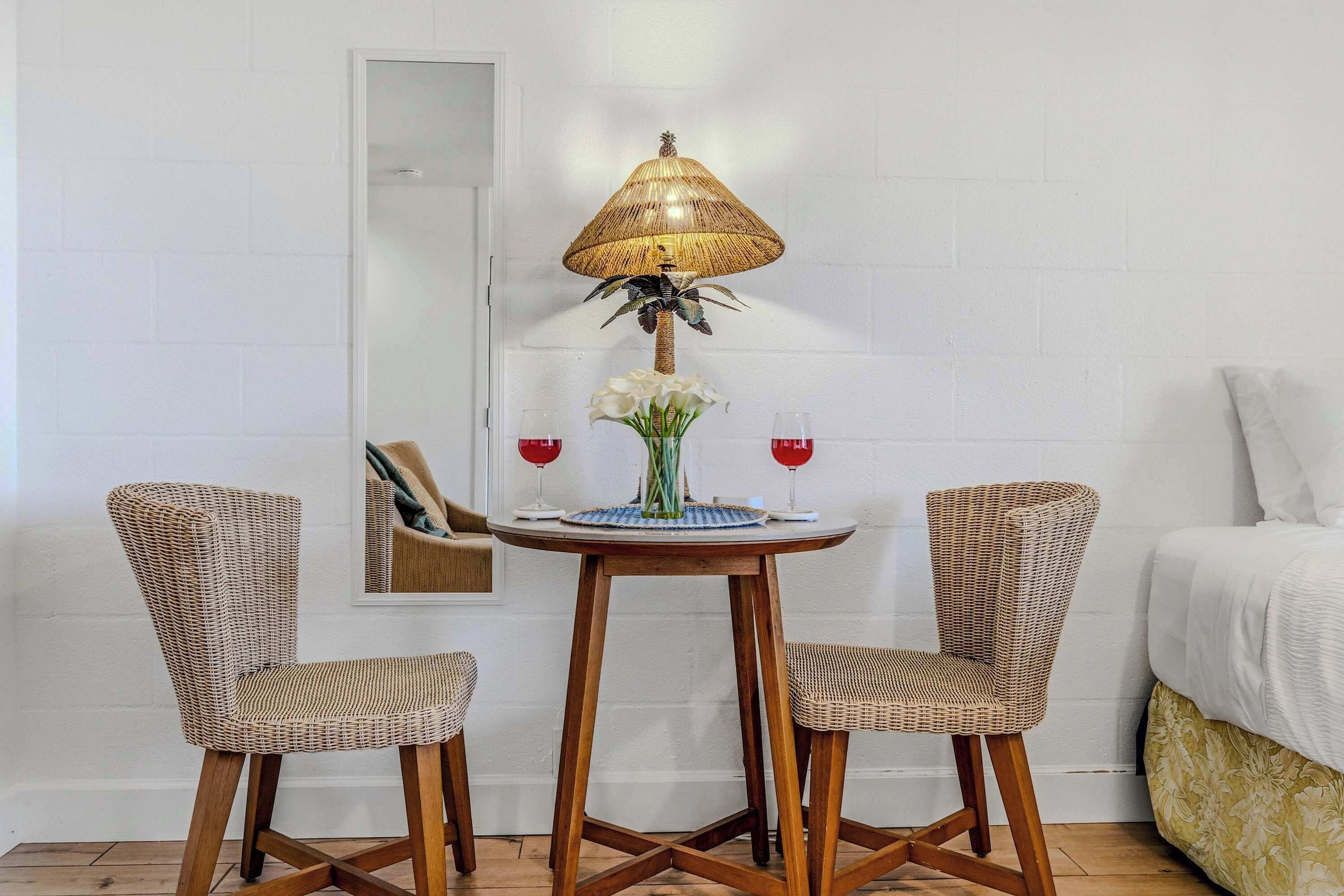 Una habitación con una mesa redonda de madera, dos sillas de mimbre, una lámpara trenzada, un jarrón con flores y dos copas de vino.