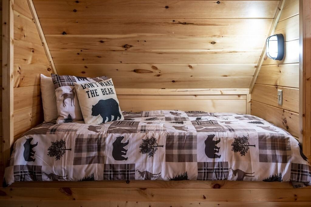 Quarto de cabana de madeira com colcha xadrez, almofada com desenho de urso e luminária fixada na parede.