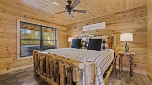 Quarto de cabana de madeira com cama, ventilador de teto, janela com persianas e um letreiro que diz 'Relaxe'.
