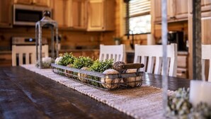 Interior em madeira com uma mesa de jantar, cadeiras, uma bandeja de metal com plantas e uma cozinha rústica ao fundo.