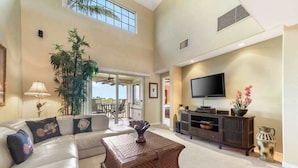 A spacious living room with a high ceiling, a large window, a sofa, a coffee table, and a TV. - *New* Fall Sale Mauna Lani Condo Near Beach Path with Waterfall Pool and Tennis (Kamuela)
