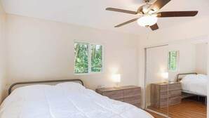 Ein Schlafzimmer mit Bett, Nachttischen, Deckenventilator, Fenster mit Blick auf Bäume und einem Spiegel.