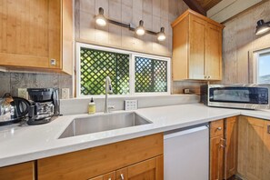 Une cuisine avec des meubles en bois, un évier en acier inoxydable, une machine à café, un four à micro-ondes et une fenêtre ornée d’un treillis décoratif vert.