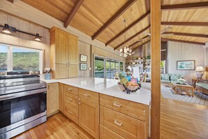 A modern kitchen with wooden cabinets, a stainless steel stove, and a large island with a bowl of fruit. - *NEW* Kealakekua Bay Oceanfront Home with Views from Every Room (Captain Cook)