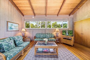 Una habitación con techo de madera, un sofá, una mesa de centro, un televisor sobre un mueble bajo y una ventana con vista a árboles.
