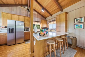 Una cocina con armarios de madera, un refrigerador de acero inoxidable, una isla central con taburetes y un ventanal con vista a un jardín.