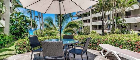 A patio with a glass table, four chairs, a lounge chair, and a large umbrella.