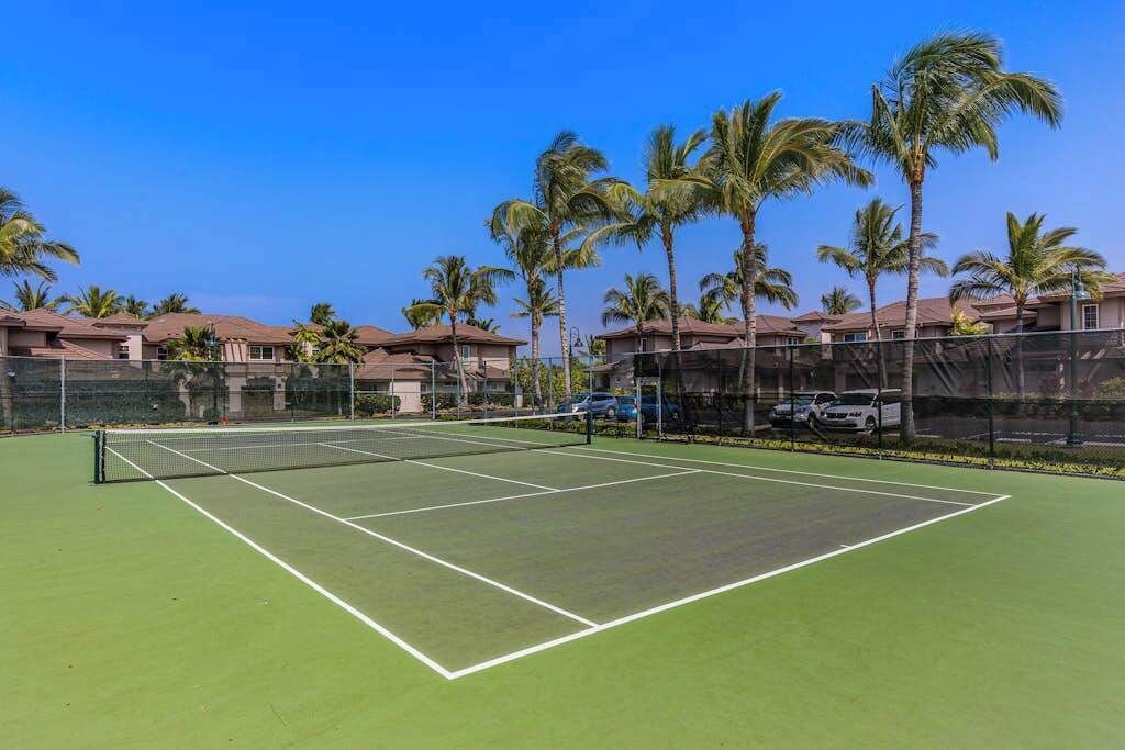 A tennis court with a net, surrounded by a fence, palm trees, and buildings in the background.