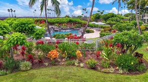 A lush garden with a pool, palm trees, and a white fence. - Waikoloa Beach Resort Villa-Golf, Pools, Hot Tub, Local Shops, Dining, Beach Fun (Waikoloa)