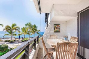 Ein Balkon mit Tisch und Stühlen, der Blick auf einen Strand mit Palmen und das Meer freigibt.