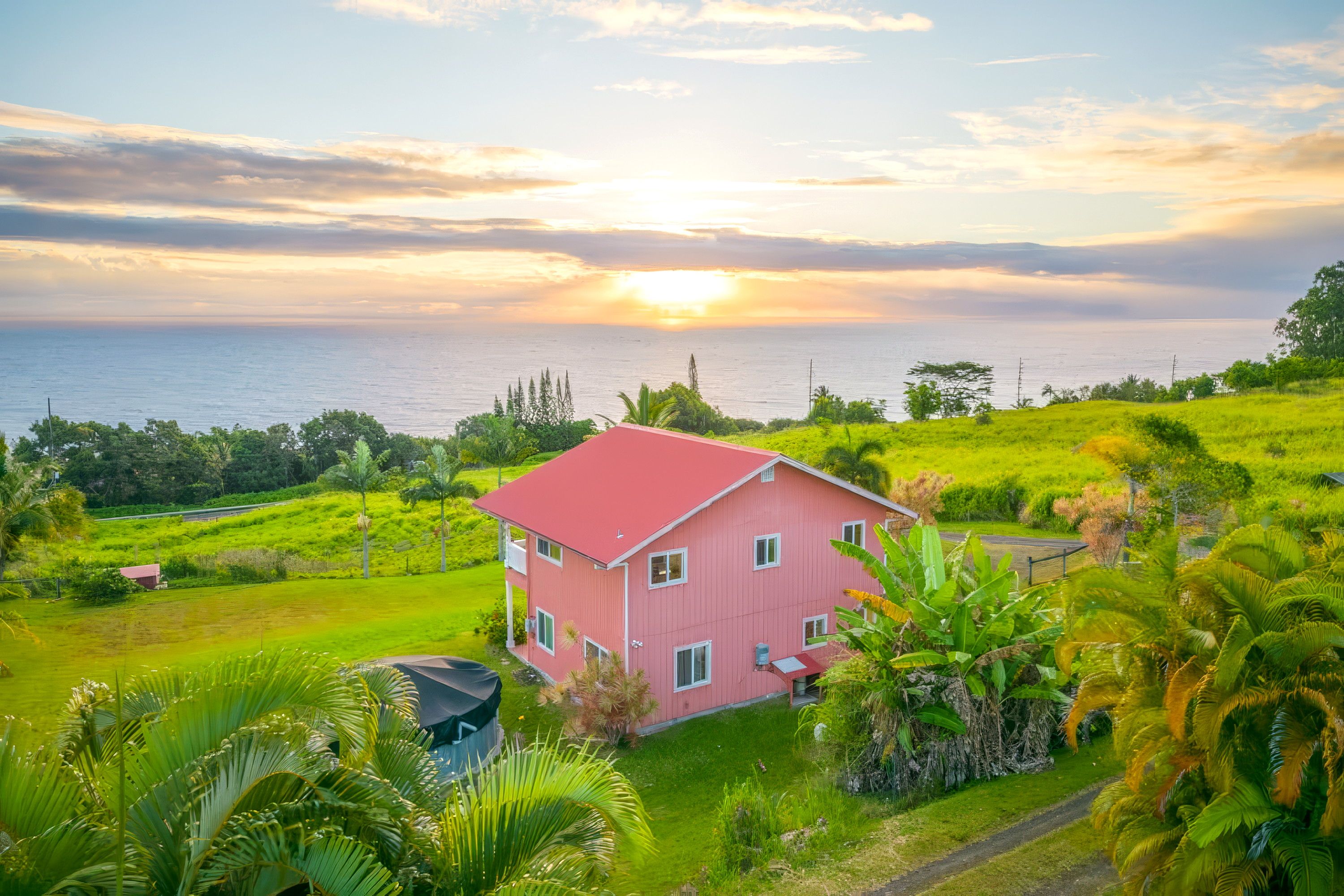 A two-story house with a red roof, surrounded by lush greenery and a view of the sea at sunset.