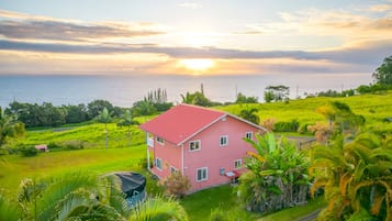 A two-story house with a red roof, surrounded by lush greenery and a view of the sea at sunset.