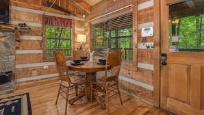 A wooden cabin interior with a dining table, chairs, a stone fireplace, and a window with blinds. - Bears in the Mist (Sevierville)