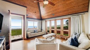 A spacious living room with a wooden ceiling, large windows, a sofa, a bench, and a television. - NEW Fall Sale Private Infinity Pool Above Kona Coffee Country (Captain Cook)