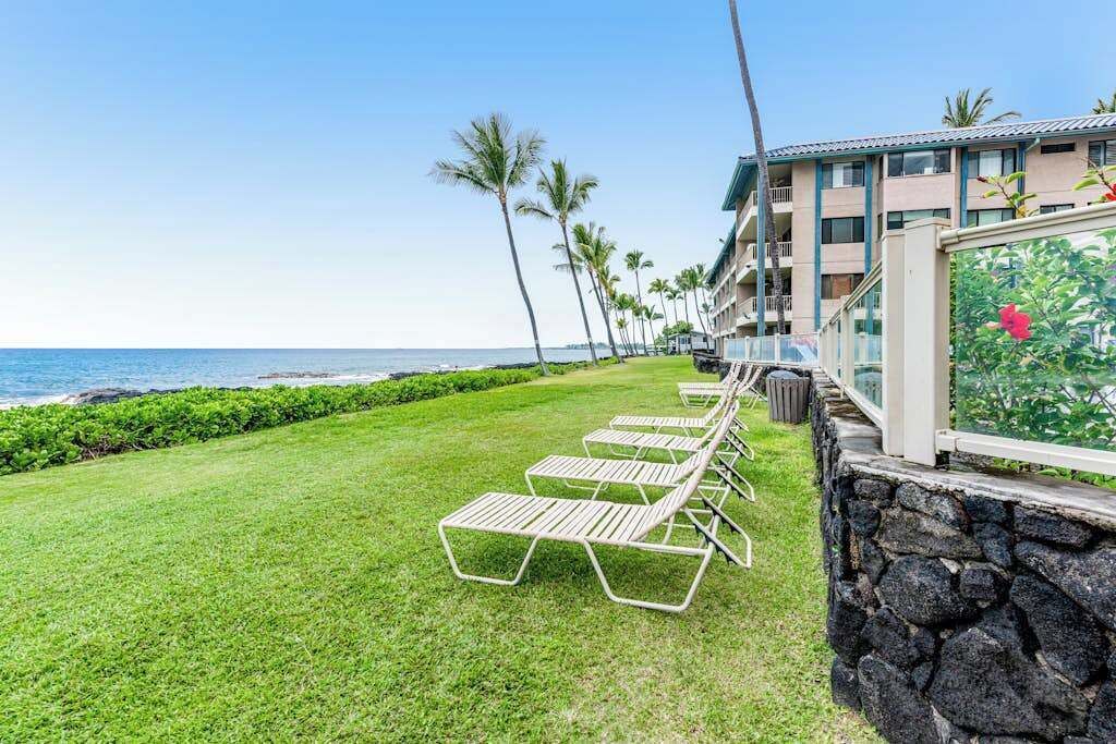 Un hôtel situé en bord de mer, avec une rangée de chaises longues, un mur en pierre et une vue sur l’océan.