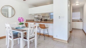 Une cuisine moderne avec des meubles blancs, un ventilateur de plafond en bois, un coin repas avec des chaises blanches et un petit bar.