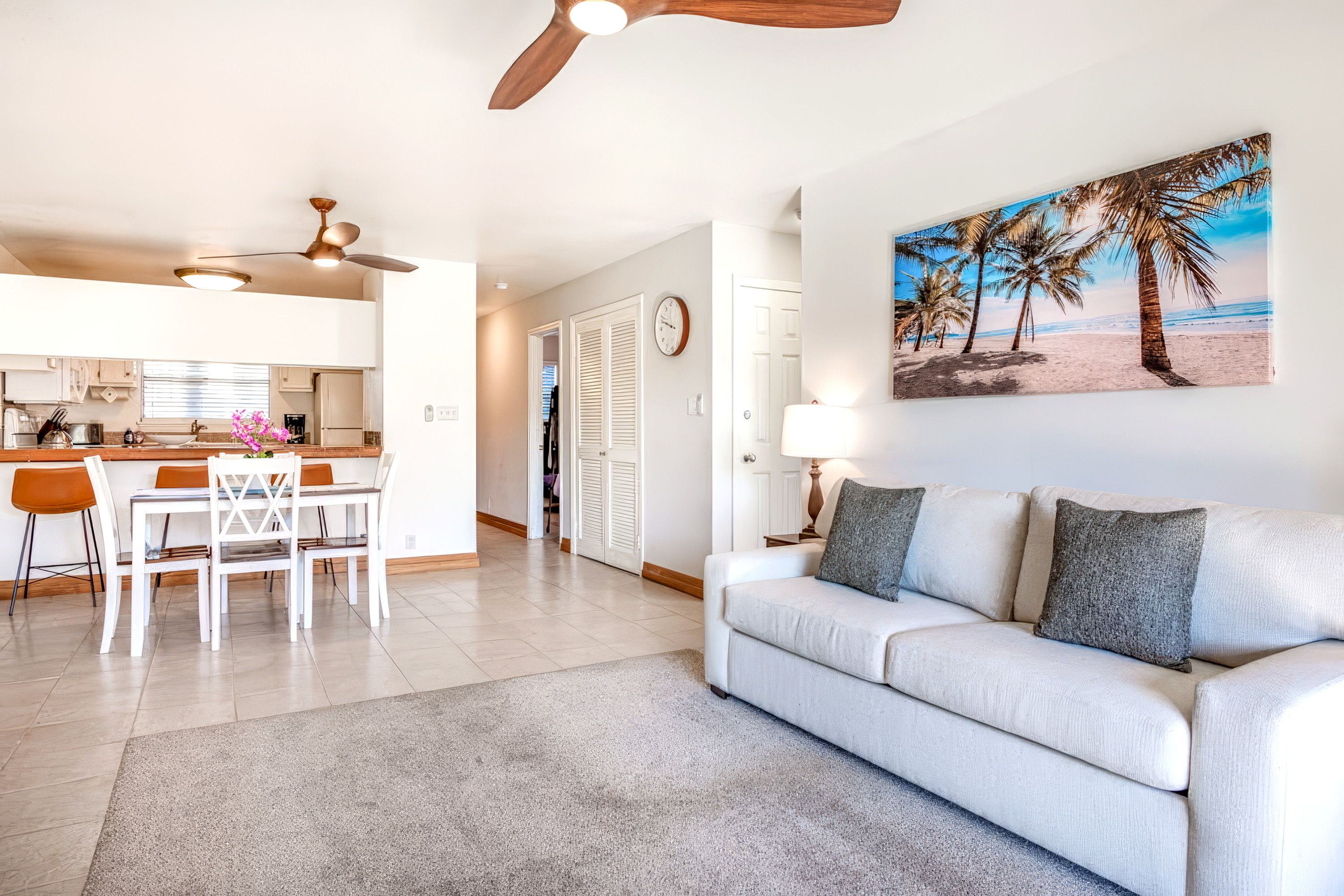 A modern living room with a sofa, a dining area with chairs, a kitchen with cabinets, a ceiling fan, and a beach-themed painting.