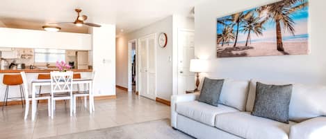 A modern living room with a sofa, a dining area with chairs, a kitchen with cabinets, a ceiling fan, and a beach-themed painting.