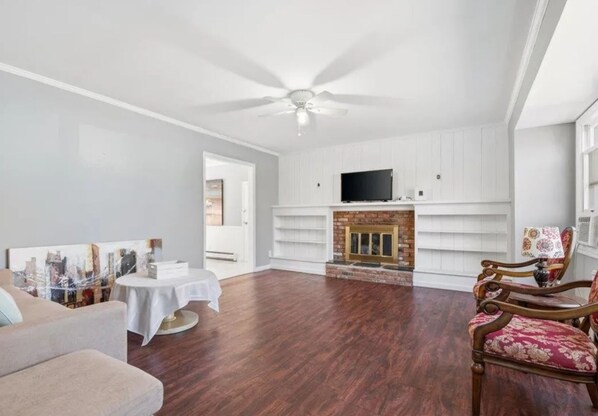 Un salon spacieux doté d’un plancher en bois franc, d’une cheminée, d’un ventilateur de plafond et d’un téléviseur.