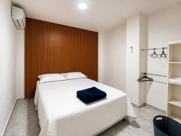 Hotellihuone, jossa on sänky, puinen pääty, valkoinen seinä, valkoinen hylly ja musta roskakori.