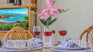 Un tavolo da pranzo con piano in vetro, due sedie, un vaso con fiori rosa e due bicchieri di vino rosso.