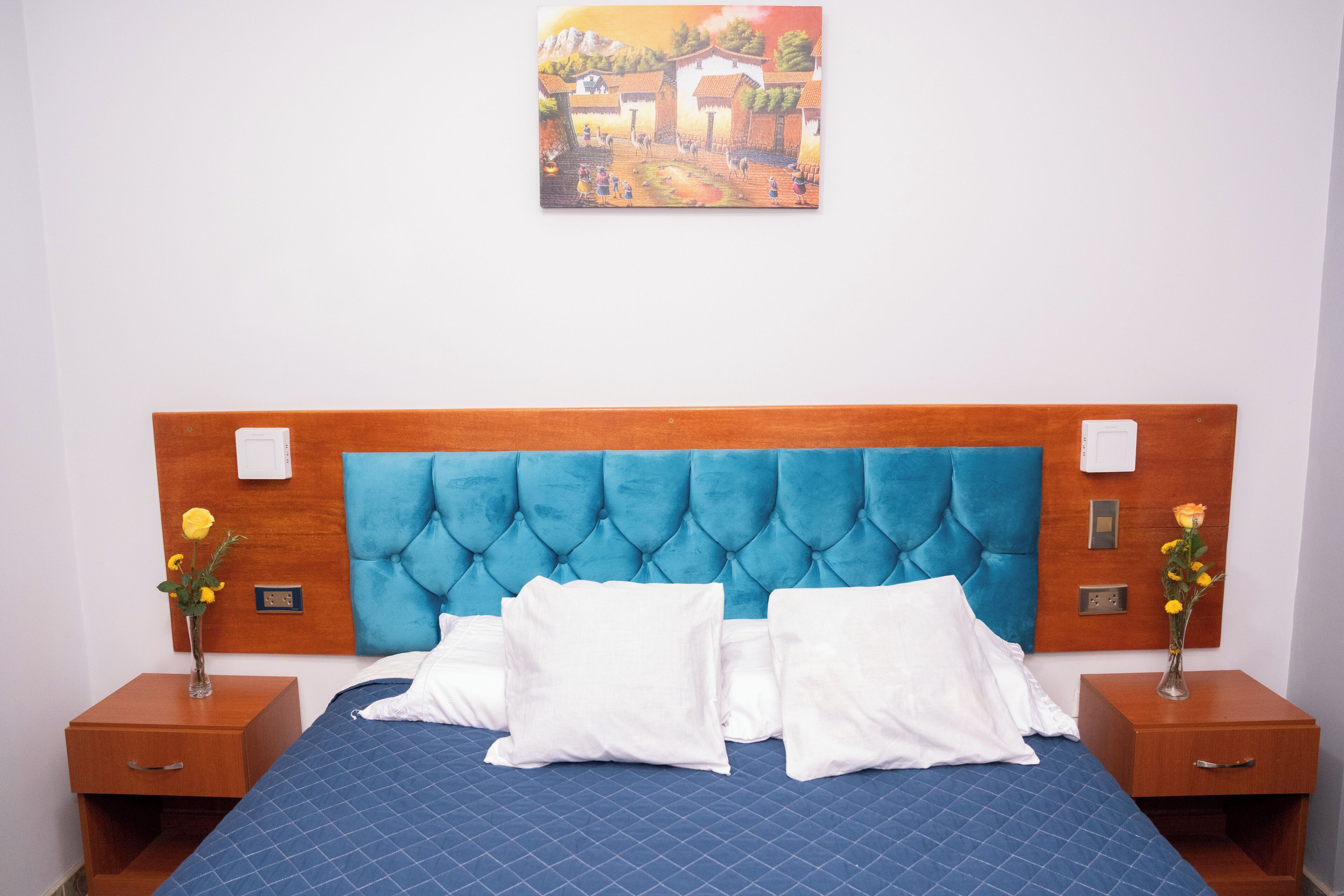 Ein Schlafzimmer mit blau gestepptem Kopfteil, weißen Kissen, einem Nachttisch aus Holz mit einer Vase voller gelber Blumen und einem Gemälde an der Wand.