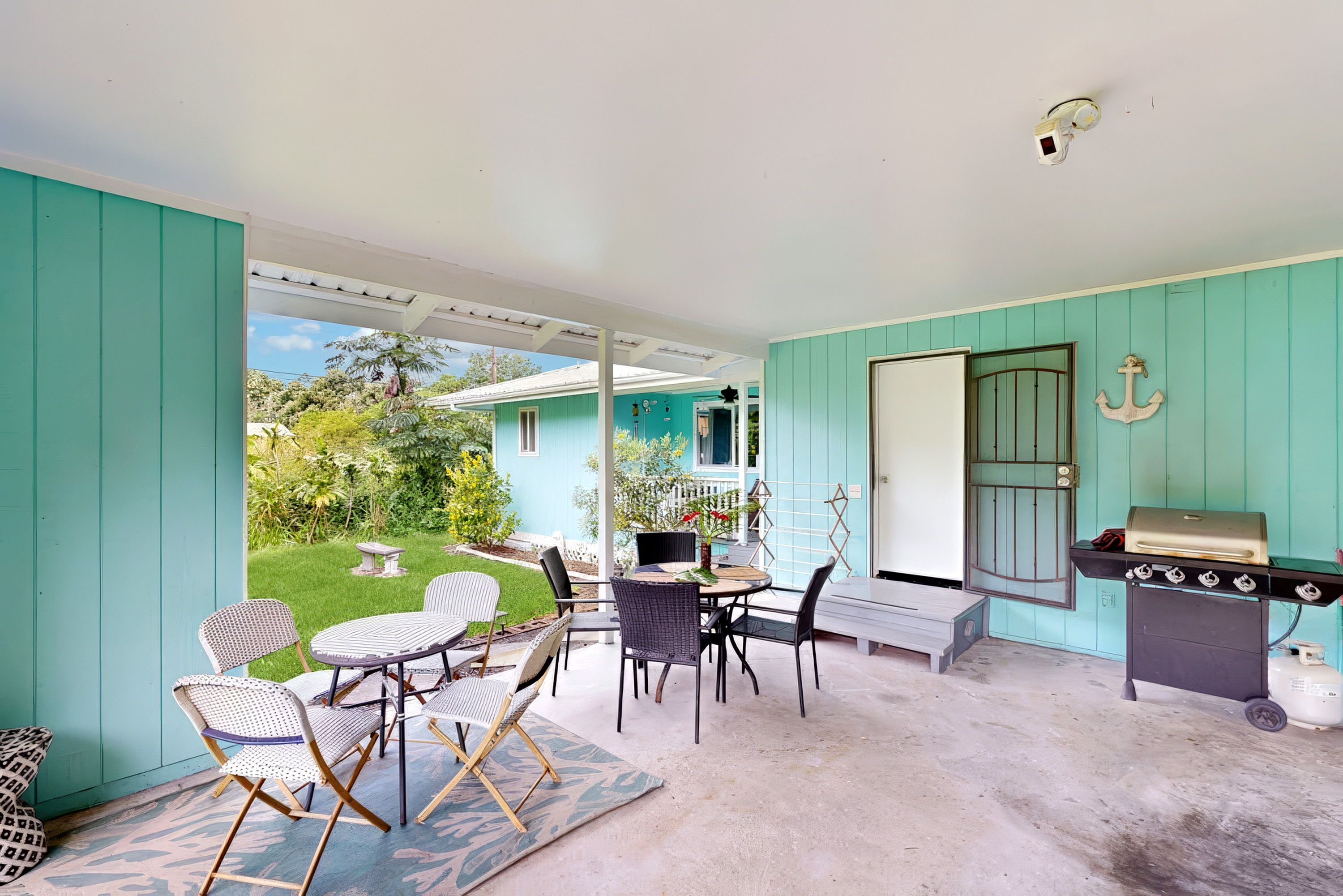A covered patio with outdoor furniture, a grill, and a view of a house with a garden.