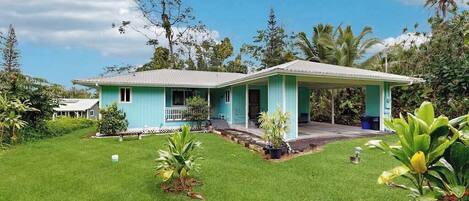 A single-story house with a covered porch, surrounded by a well-maintained lawn and tropical vegetation.